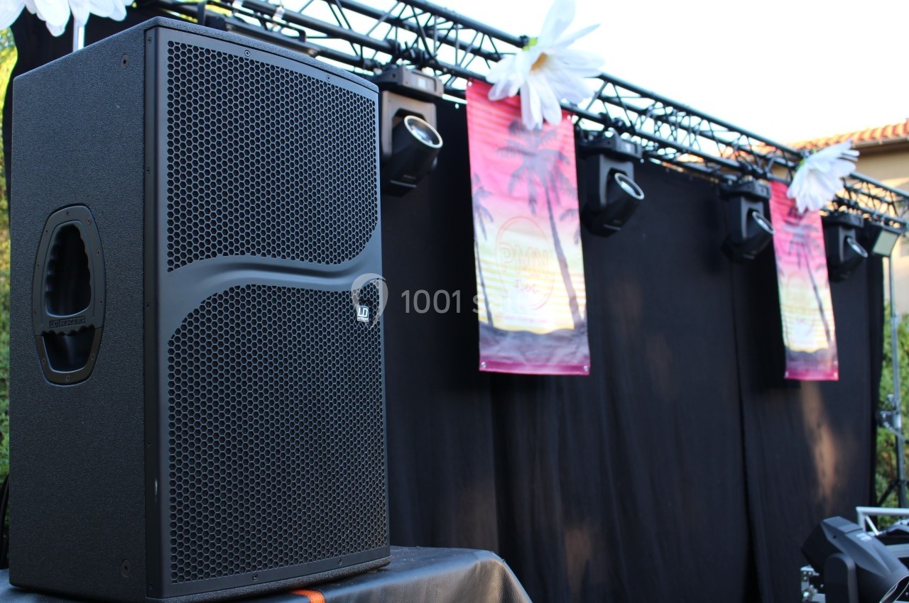 Enceinte audio noire posée devant une scène décorée de fleurs blanches et de bannières suspendues.