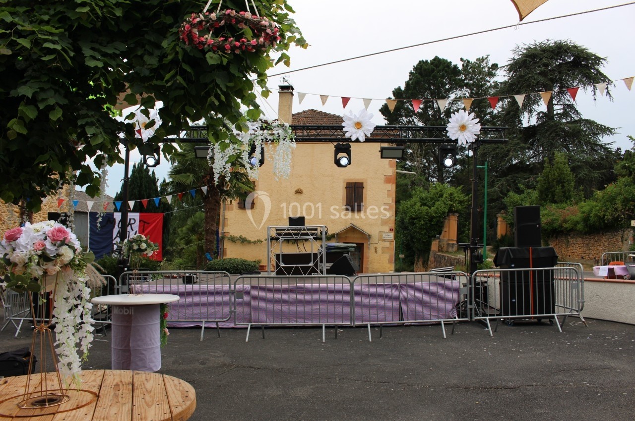 Place décorée pour un événement avec guirlandes, fleurs suspendues, scène et barrières devant un bâtiment en pierre.