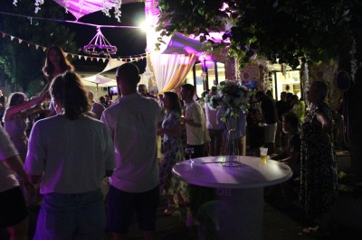 Personnes dansant et discutant lors d'une soirée en extérieur, éclairée par des lumières colorées et décorée de guirlandes.