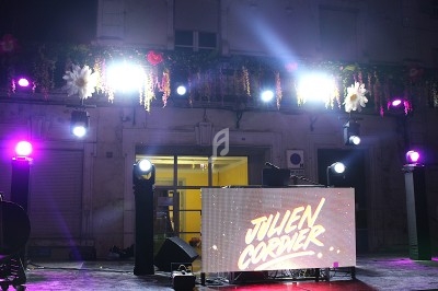 Scène de DJ en extérieur avec éclairage coloré, écran affichant un nom et décorations florales suspendues.