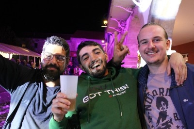Trois hommes souriants posent ensemble lors d'un événement nocturne avec des lumières colorées en arrière-plan.