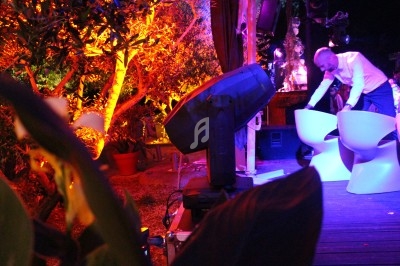 Un homme ajuste des chaises blanches sur une terrasse éclairée par des lumières colorées dans un cadre extérieur.