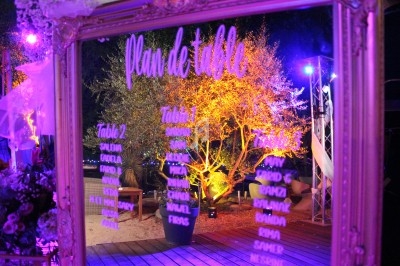 Plan de table écrit sur un miroir doré, reflétant un décor extérieur illuminé avec des arbres et des lumières colorées.