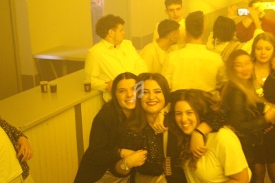 Trois jeunes femmes souriantes posent ensemble dans une ambiance festive et lumineuse, entourées d'autres personnes.