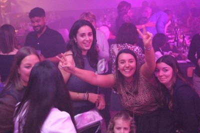 Des personnes souriantes dans une ambiance festive et lumineuse, entourées de convives à table.