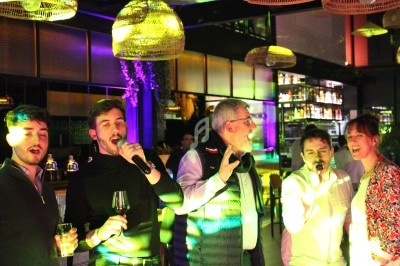 Des personnes chantent et s'amusent dans un bar éclairé par des lumières colorées, avec un décor moderne en arrière-plan.