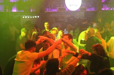 Personnes dansant en cercle dans une ambiance festive avec éclairage jaune et violet dans un bar animé.