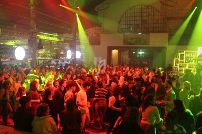 Foule dans une salle éclairée par des lumières rouges et vertes, avec un bar en arrière-plan.