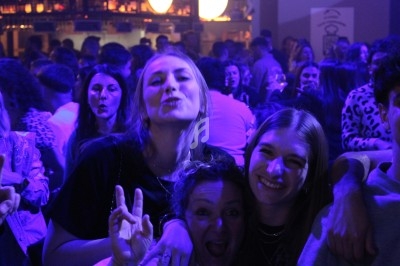 Trois femmes souriantes posent ensemble dans une ambiance festive et éclairée par des lumières bleutées.