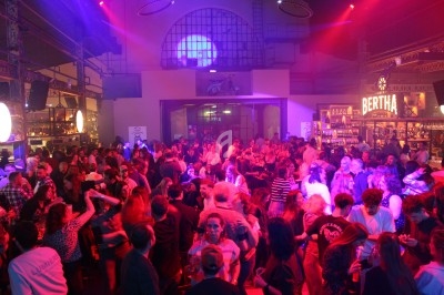 Foule dans une salle animée avec éclairage coloré, bar en arrière-plan et ambiance festive.