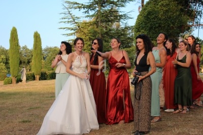 Un groupe de femmes élégamment vêtues, dont une en robe de mariée, riant dans un jardin ensoleillé.