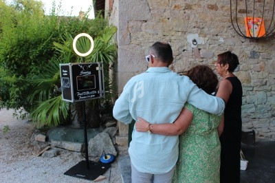 Trois personnes posent devant une borne photo en extérieur, près d'un mur en pierre et de végétation.