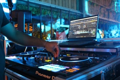 Un DJ manipule une table de mixage éclairée, avec un ordinateur affichant un logiciel de musique en arrière-plan.