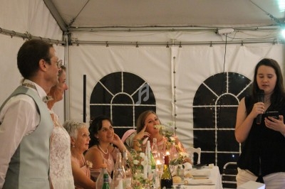 Une femme lit un discours au micro devant des invités assis à une table décorée dans une tente éclairée.