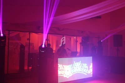 Salle de fête avec éclairage violet, DJ derrière une table lumineuse et décorations suspendues au plafond.