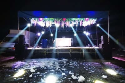 Scène extérieure illuminée de nuit avec jeux de lumière, décoration florale suspendue et mousse au sol.