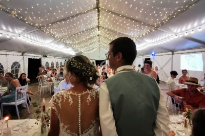 Un couple de mariés de dos dans une salle de réception décorée de guirlandes lumineuses, entouré d'invités.