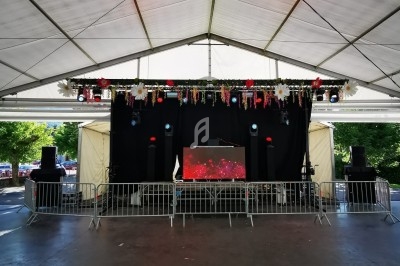 Scène couverte en plein air avec équipement sonore, décorations florales suspendues et barrières de sécurité.