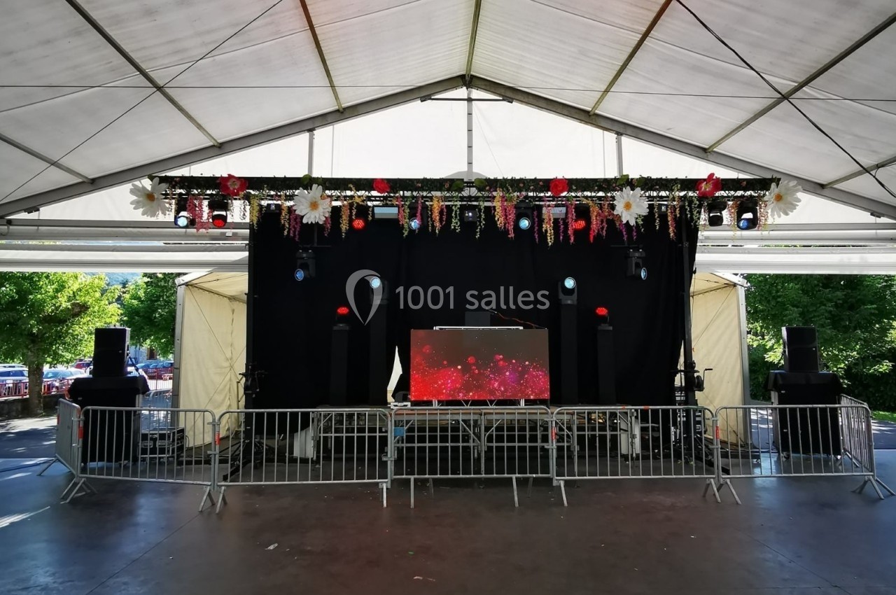 Scène couverte avec une table de mixage, des enceintes et une décoration florale, entourée de barrières de sécurité.