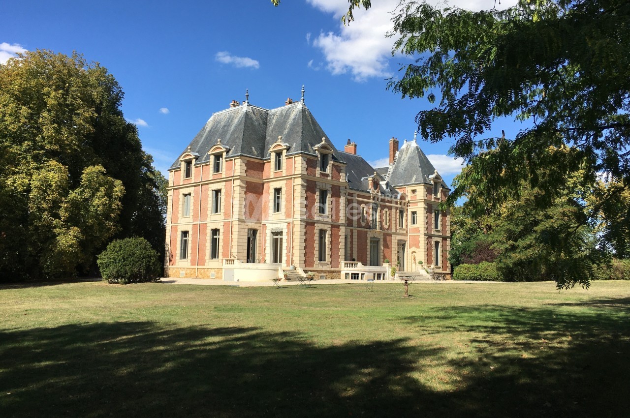 Château en briques et pierres entouré d'un parc arboré sous un ciel bleu.