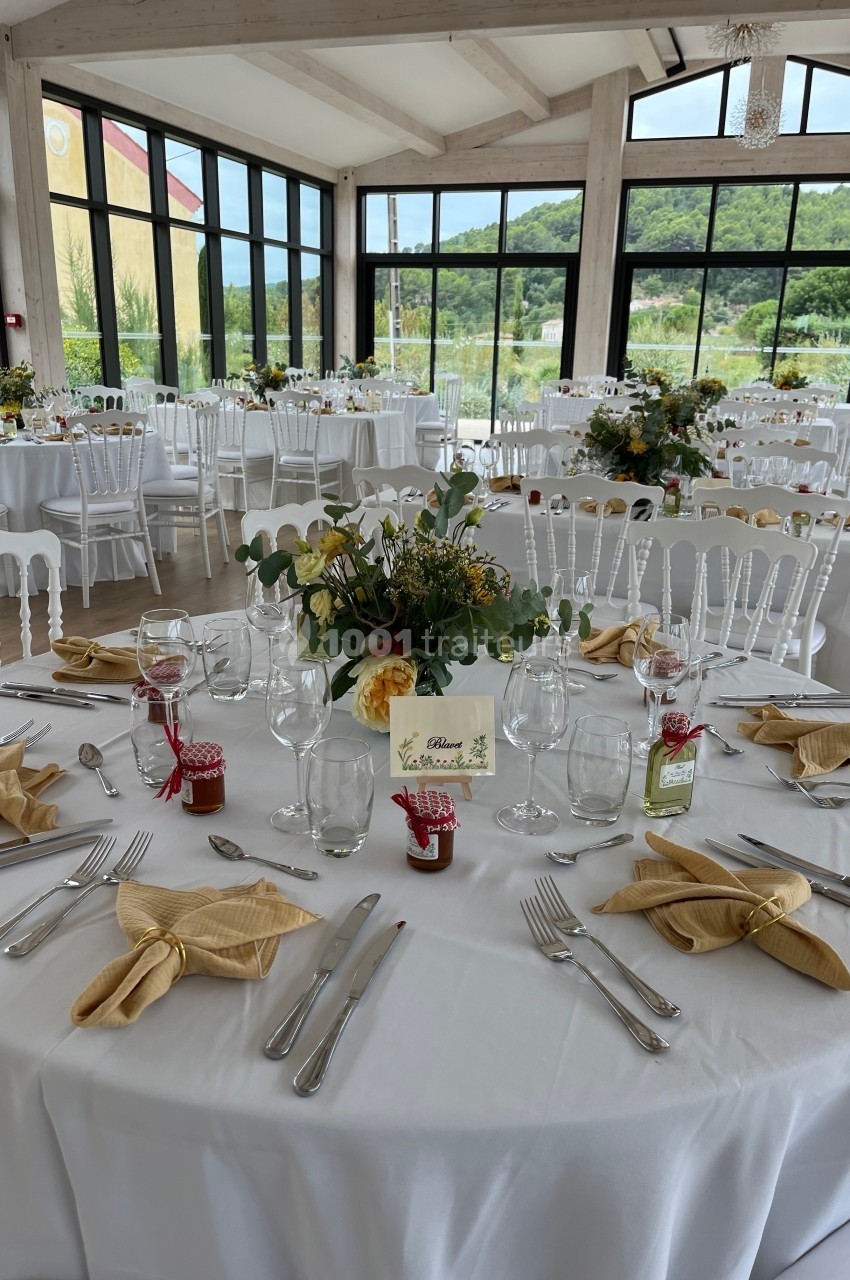 Salle de réception lumineuse avec tables rondes dressées, nappes blanches, décorations florales et couverts élégants.