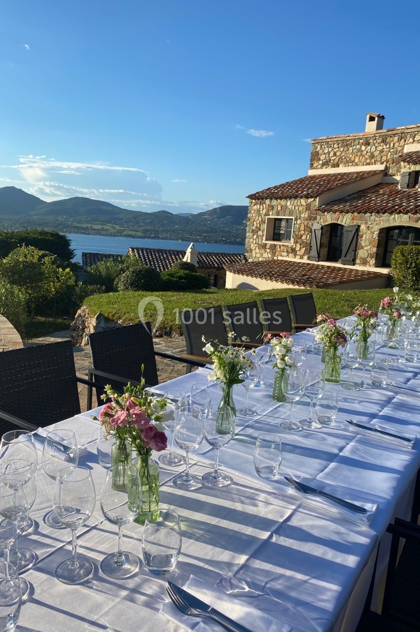 Table dressée en extérieur avec nappes blanches, verres et fleurs, devant une maison en pierre et un paysage de montagne.
