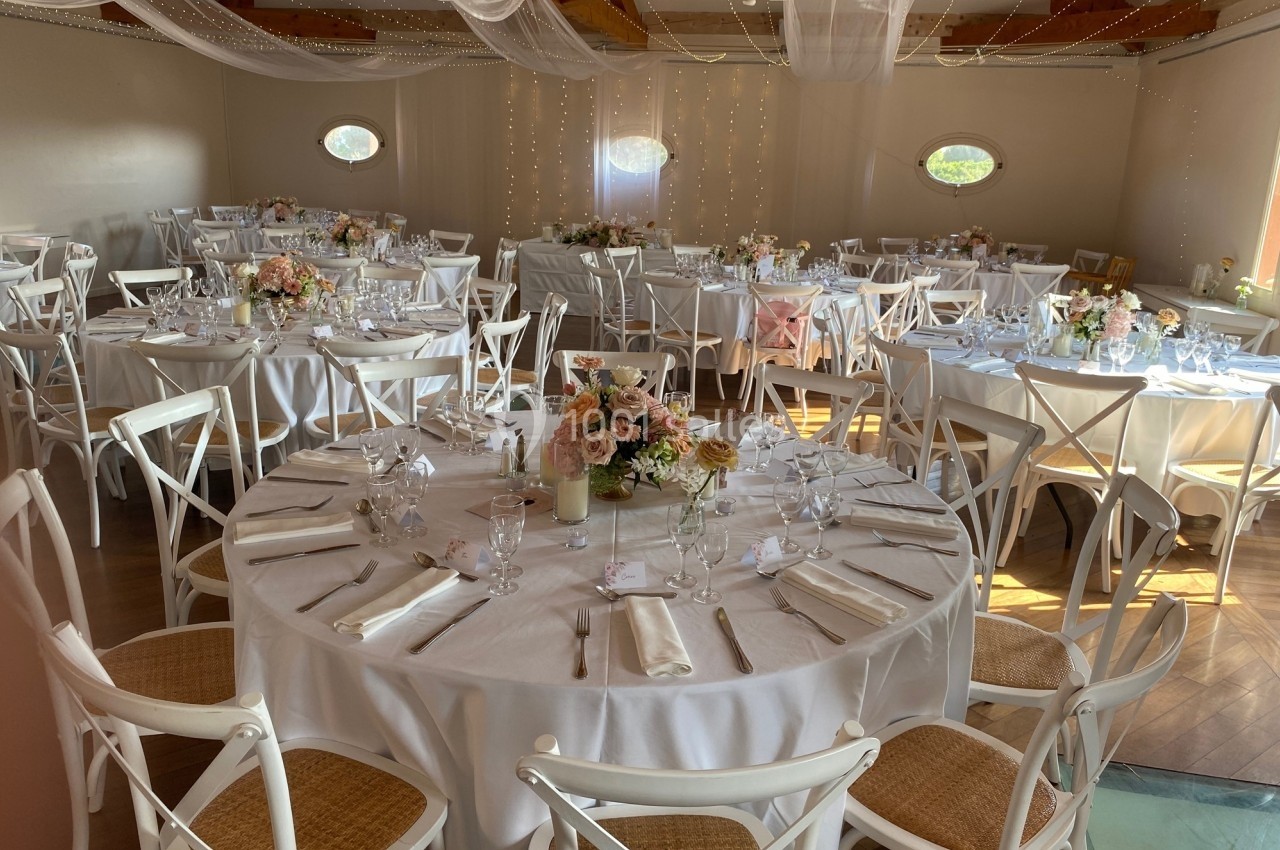 Salle de réception décorée pour un mariage, avec tables rondes dressées, nappes blanches et arrangements floraux.