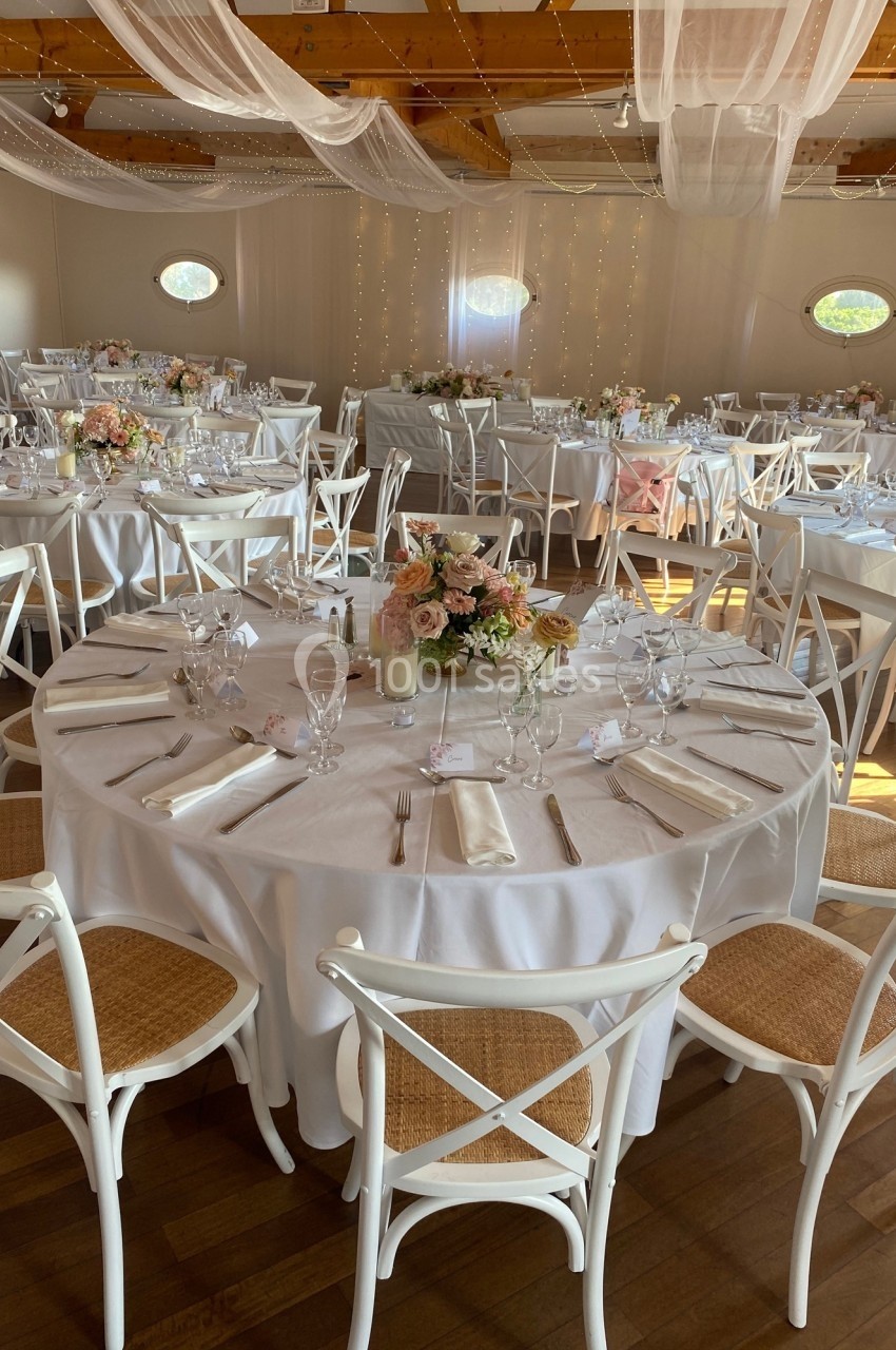Salle décorée pour un événement avec des tables rondes dressées, nappes blanches, chaises en bois et fleurs au centre.