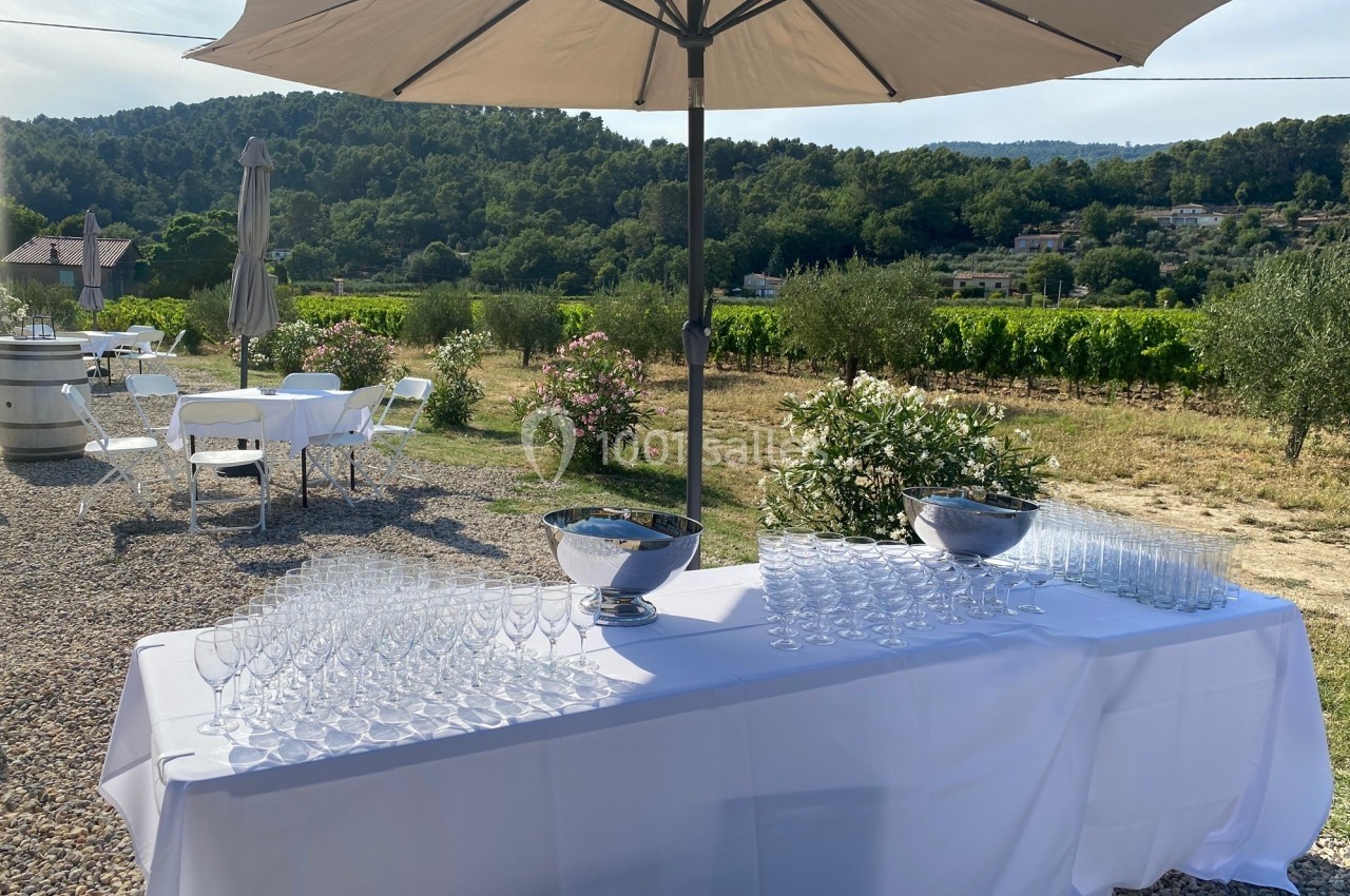 Table dressée avec des verres à pied sous un parasol, en extérieur, avec vue sur des vignes et des collines verdoyantes.