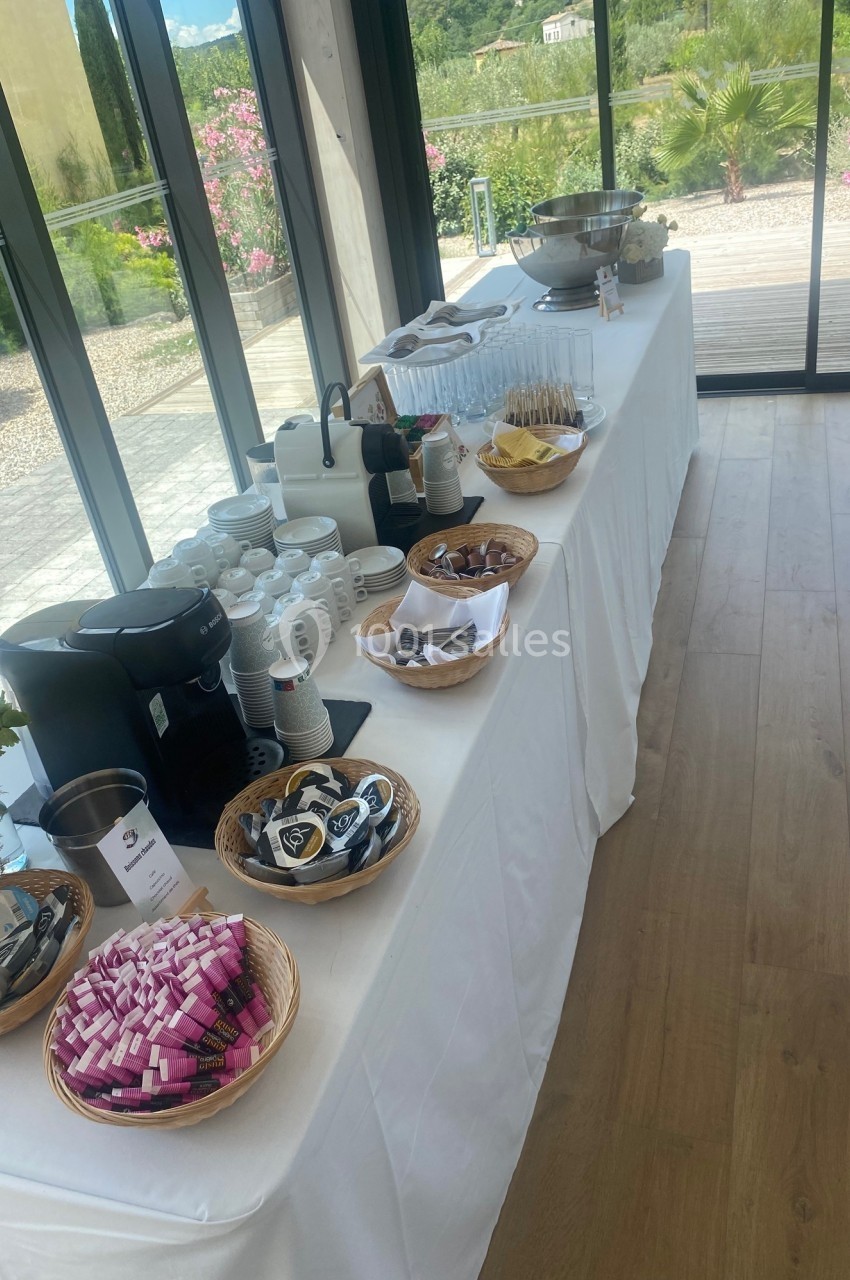 Table dressée avec boissons chaudes, viennoiseries et condiments, située près de grandes baies vitrées donnant sur un jardin.