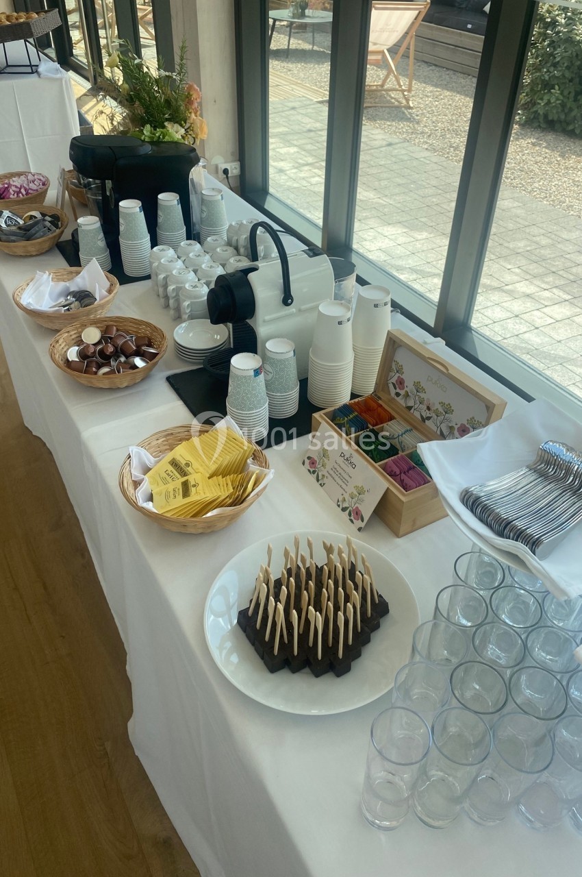 Buffet avec boissons chaudes, gobelets, couverts, chocolats, sachets de thé et mignardises sur une table blanche.