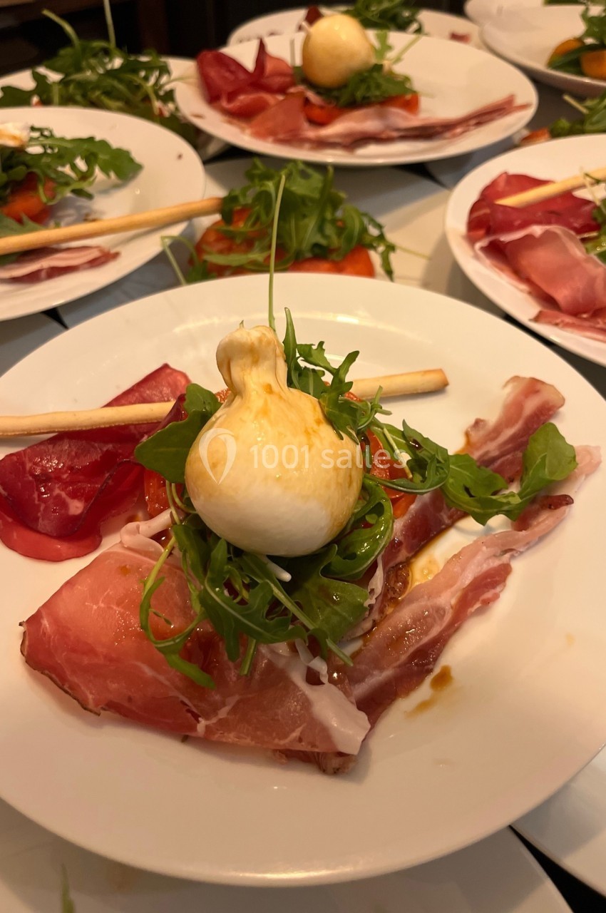 Assiette composée de jambon cru, roquette, tomate, burrata et gressin, présentée sur une table.