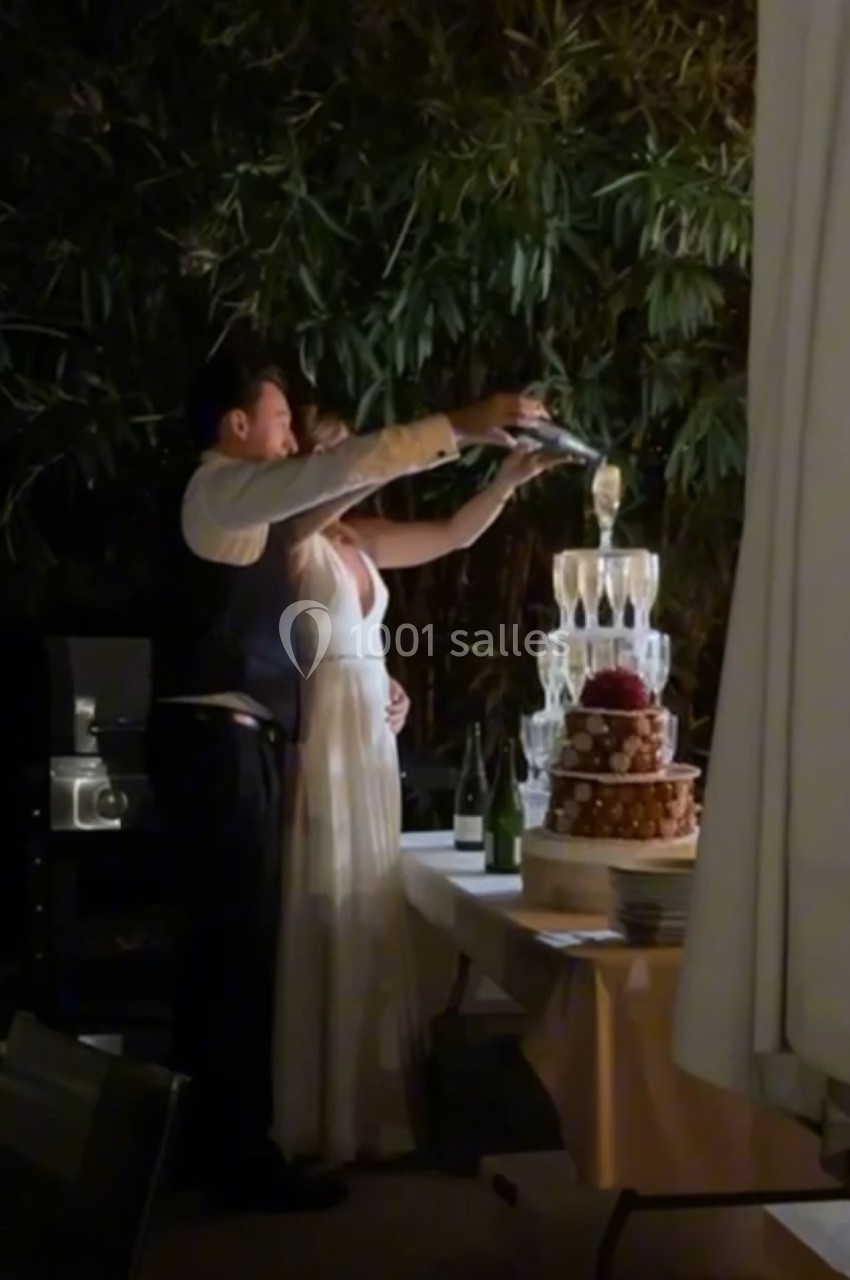 Un couple verse du champagne dans une pyramide de verres lors d'une réception en extérieur, entouré de végétation.