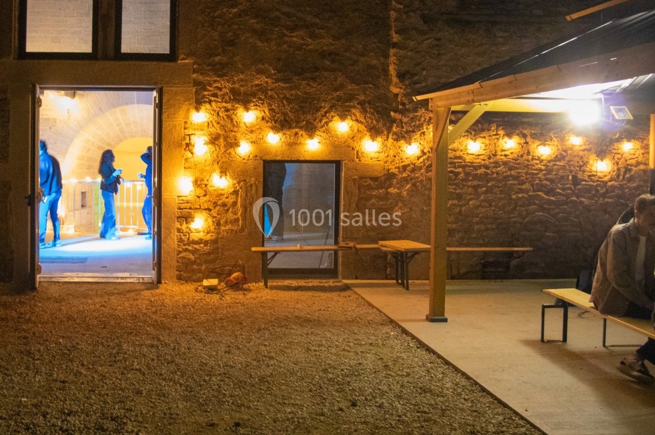 Cour extérieure éclairée par des guirlandes lumineuses, avec une porte ouverte révélant une pièce illuminée en bleu.