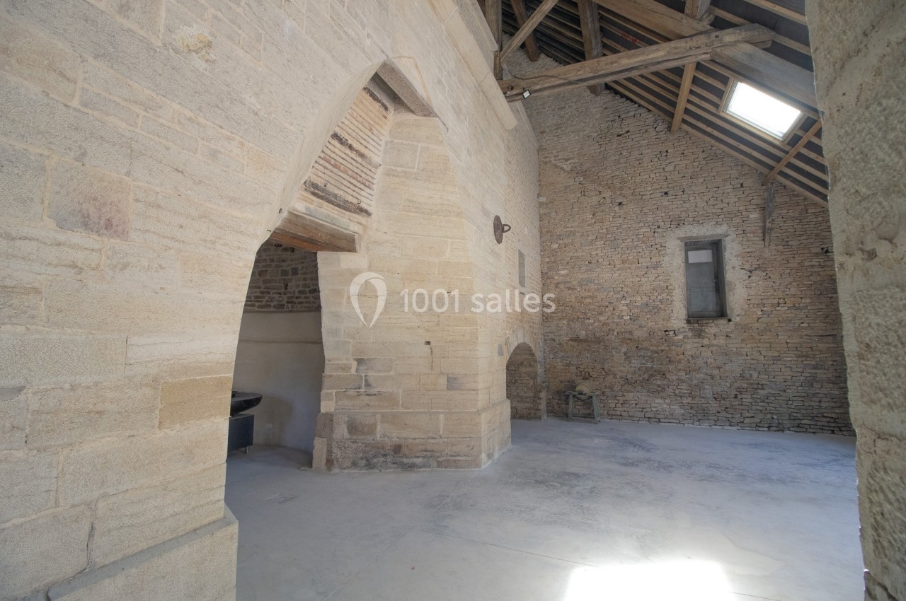 Intérieur d'un bâtiment en pierre avec des arches, un sol en béton et une charpente en bois visible sous le toit.