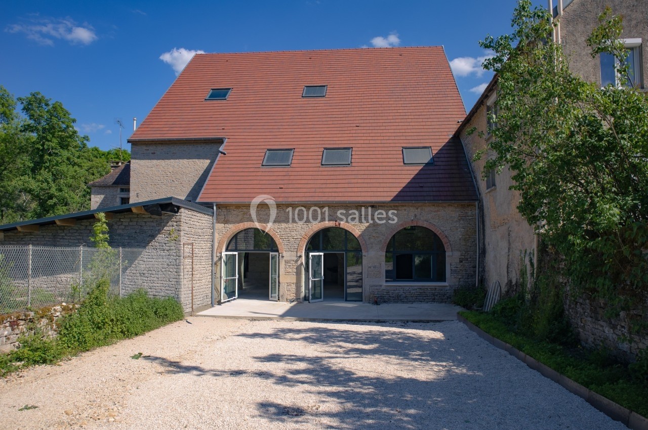 Façade d'un bâtiment rénové en pierre avec toit en tuiles, grandes arches vitrées et cour en gravier.