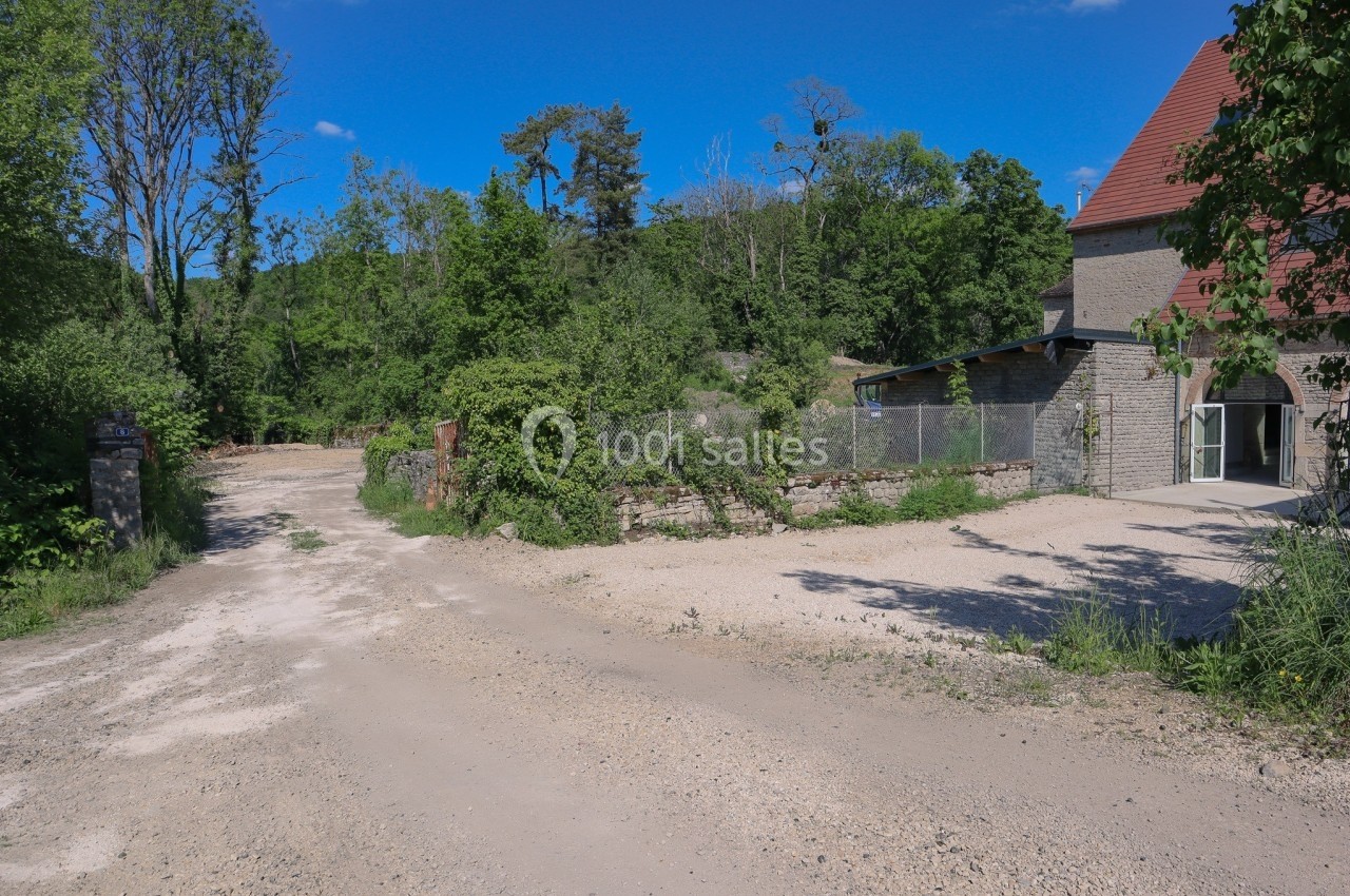 Chemin de terre bordé de végétation menant à un bâtiment en pierre avec un toit rouge, sous un ciel dégagé.