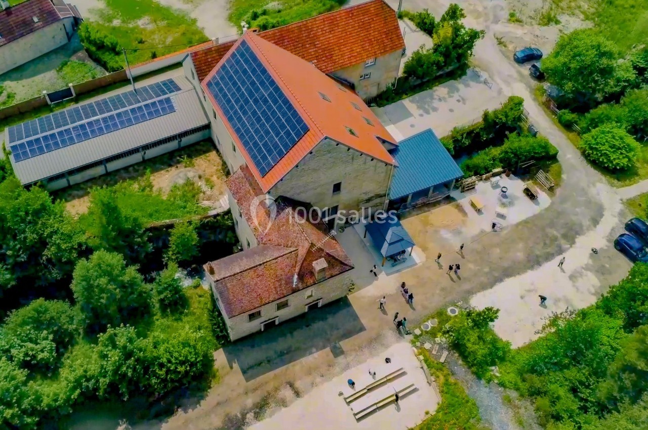 Vue aérienne d'un bâtiment en pierre avec toit en tuiles et panneaux solaires, entouré de verdure et d'une cour.