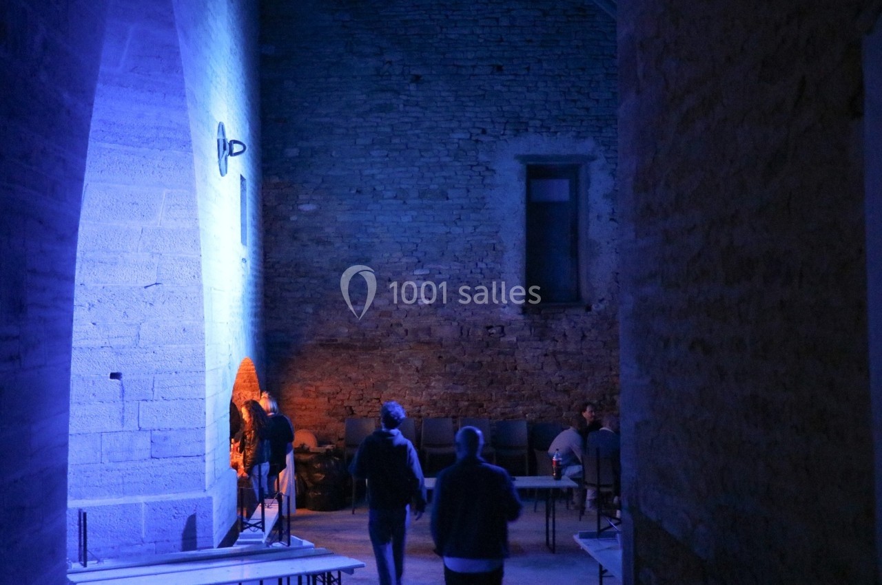 Personnes marchant dans un couloir en pierre éclairé par une lumière bleue, avec des chaises alignées au fond.