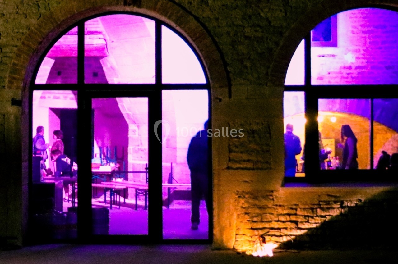Vue nocturne d'une façade en pierre avec des arches vitrées, révélant un intérieur éclairé en violet et des silhouettes.