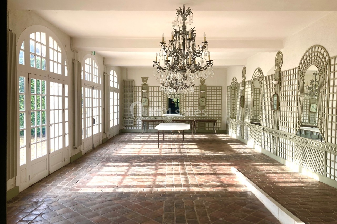 Salle lumineuse avec sol en carreaux de terre cuite, grandes fenêtres, miroirs muraux et lustre central en cristal.