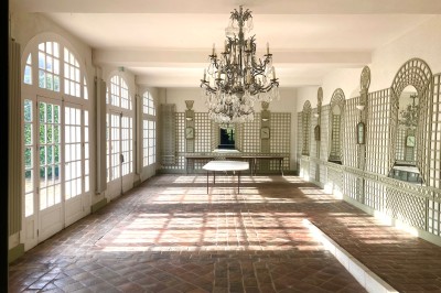 Cuisine ancienne avec une grande table en bois, un sol en pierre, des fenêtres lumineuses et des éléments rustiques.