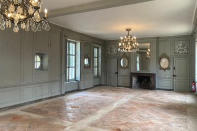 Cuisine ancienne avec une grande table en bois, un sol en pierre, des fenêtres lumineuses et des éléments rustiques.