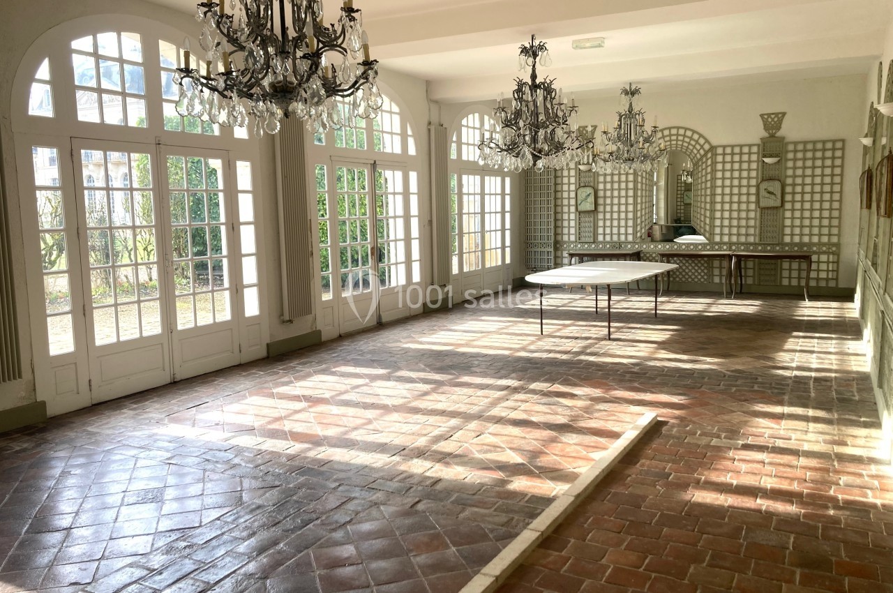 Salle lumineuse avec sol en carreaux de terre cuite, grandes fenêtres cintrées, miroirs et lustres en cristal.