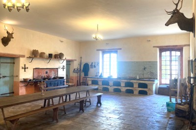 Cuisine ancienne avec une grande table en bois, un sol en pierre, des fenêtres lumineuses et des éléments rustiques.