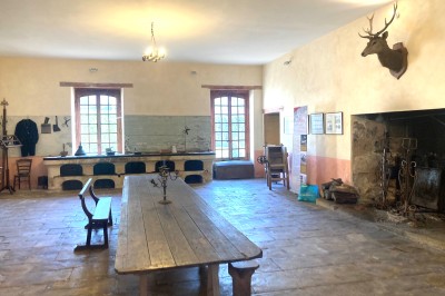 Cuisine ancienne avec une grande table en bois, un sol en pierre, des fenêtres lumineuses et des éléments rustiques.