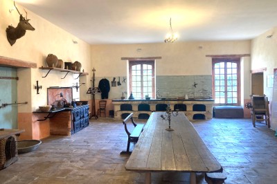Cuisine ancienne avec une grande table en bois, un sol en pierre, des fenêtres lumineuses et des éléments rustiques.