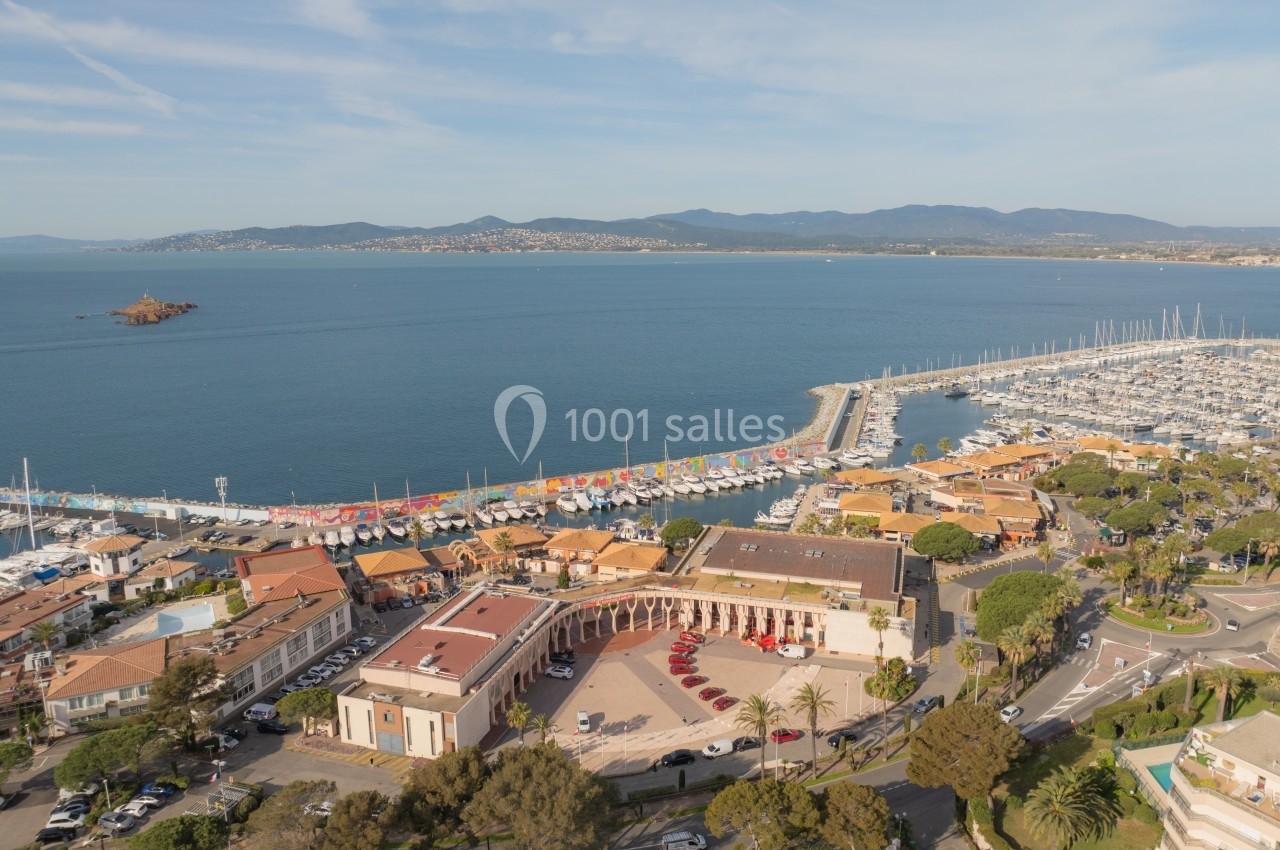 Vue aérienne d'un port de plaisance bordé de bâtiments et d'une promenade, avec la mer et des collines en arrière-plan.
