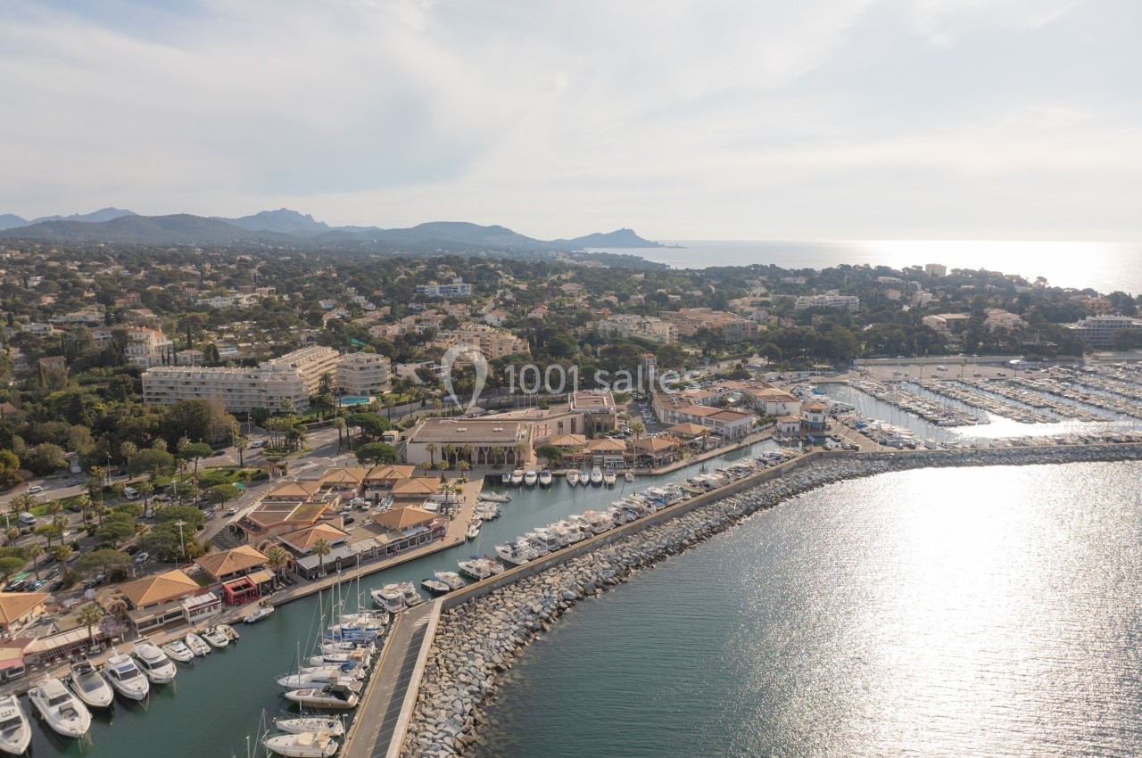 Vue aérienne d'un port de plaisance avec des bateaux amarrés, entouré de bâtiments et de collines en arrière-plan.