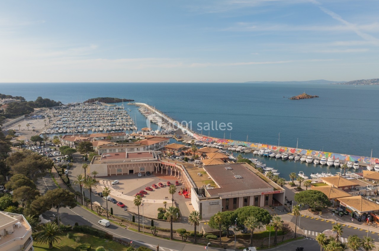 Vue aérienne d'un port de plaisance avec des bateaux amarrés, bâtiments environnants et mer en arrière-plan.